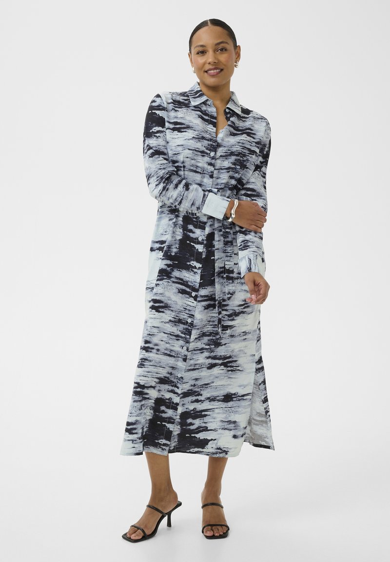 Femme portant une robe longue boutonnée à motifs abstraits noir et blanc avec manches longues et sandales à talons noires à lanières.