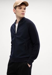 Mornarska zip-up jakna z teksturiranimi paneli na ramenih, z zaključenim žepom in rebrastimi manšetami. Nosi se čez belo srajco, v kombinaciji z črnimi hlačami.