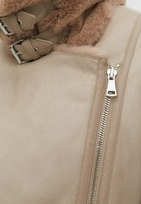 Beige faux suede jacket with a soft, furry collar, silver zipper, and two adjustable strap closures near the neckline.