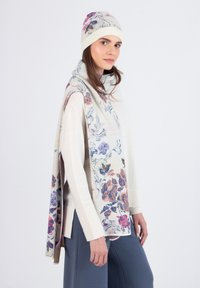Floral-patterned cream scarf drapes over a light sweater; matching cream beanie with ribbed cuff. Soft texture, warm, and cozy appearance.