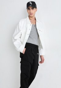 Weiße Jeansjacke über einem grauen Tanktop, kombiniert mit schwarzen Cargo-Hosen. Mit Taschen, Gummizugbund und einer schwarzen Mütze.