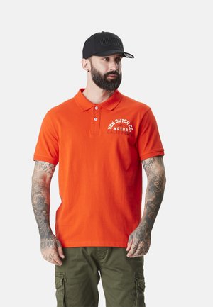 Homme barbu avec tatouages portant une casquette noire, un polo orange avec le texte "Von Dutch Co.", et un pantalon cargo vert olive, regardant sur le côté.