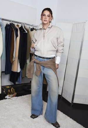 Kvinne iført beige jakke med glidelås og vide, falmede jeans står i rom med klesstativ og sko på teppet gulv.