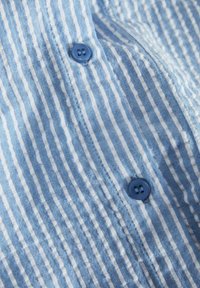 Ριγέ πουκάμισο σε ανοιχτό μπλε και λευκό, με δύο μπλε κουμπιά. Το ύφασμα έχει μαλακή υφή με κατακόρυφες γραμμές.