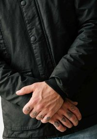 Giacca nera e tessurizzata con bottoni a pressione; mani incrociate che mostrano tatuaggi geometrici sulle dita. Tessuto morbido, polsini aderenti.