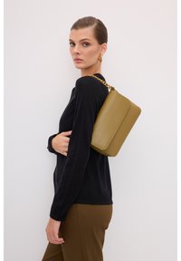 Olijfgroene leren handtas met gouden kettingriem, rechthoekige vorm en gestikte accenten, gedragen over een zwarte top door een model.