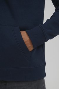 Navy blue sweatshirt featuring a kangaroo pocket, ribbed cuffs, and a textured fabric. Close-up showing hand resting in the pocket.