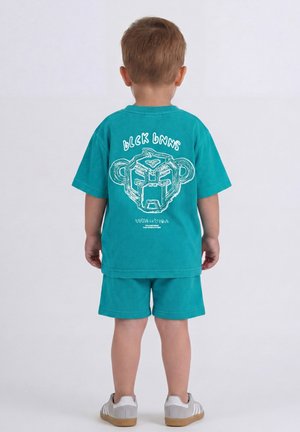 Jong kind met turquoise shirt en short met een gestileerd apengezicht en tekst op de rug, staat met de rug naar voren tegen een effen achtergrond.