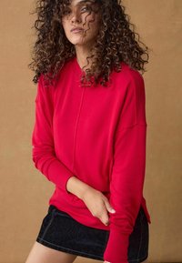 Felpa rossa con polsini e orlo a costine, con una vestibilità rilassata. Abbinata a una mini gonna in denim scuro su uno sfondo neutro.