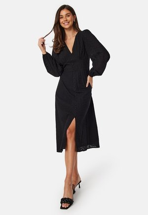 Zwarte jurk met lange mouwen, V-hals, knopen aan de voorkant en een hoge split tot aan de dij; textuurstof en een relaxte silhouet.