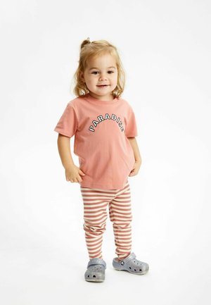 Rosa Kurzarm-T-Shirt mit dem Text "PARADISE", kombiniert mit gerippten, gestreiften Leggings in Pfirsich und Weiß sowie grauen Slip-On-Schuhen.