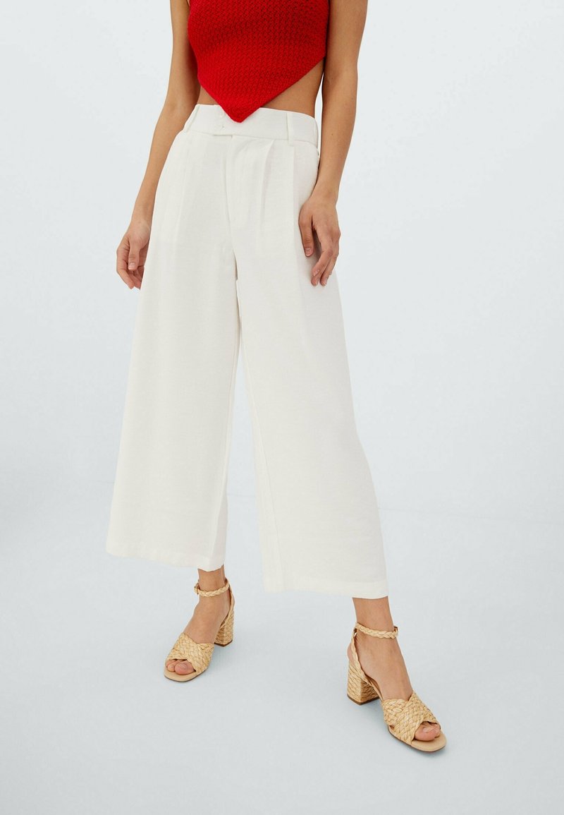 Pantalon blanc à jambes larges avec plis sur le devant et taille haute, associé à des sandales tressées à talons couleur tan. Fond neutre.