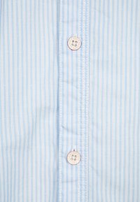 Ljusblå skjorta med knappar och vertikala vita ränder, med sex knappar och synliga sömmar längs knappslån.