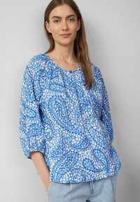 Blaue Bluse mit Muster, dreiviertel Ärmel, runder Ausschnitt und gerafften Details; verfügt über eine Mischung aus hellblauen und dunkelblauen Formen auf weißem Hintergrund.
