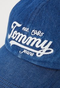 Blue denim cap featuring white embroidered "Tommy Jeans" logo with established year 1985. Curved brim and textured fabric.