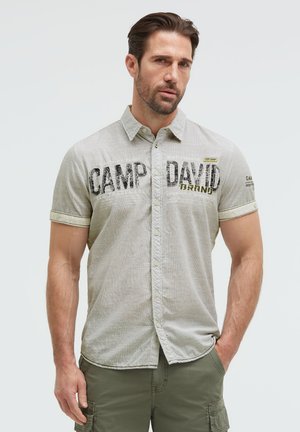Mann trägt ein hellgraues kurzärmeliges Hemd mit dem Text "Camp David" und olivgrüne Cargohosen, steht mit einer Hand in der Tasche.