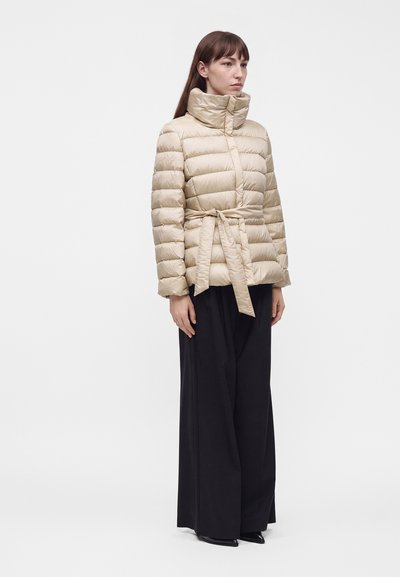 Femme aux cheveux bruns et lisses portant une doudoune beige ceinturée et un pantalon large noir, debout sur un fond blanc.