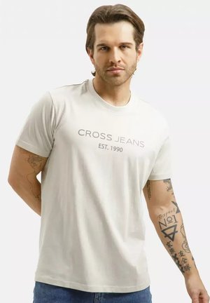 Mężczyzna o średnio brązowych włosach, ubrany w białą koszulkę "CROSS JEANS EST. 1990" i niebieskie jeansy, pokazujący tatuaże na obu ramionach.