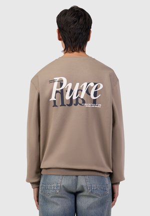 Man met kort donker haar die een bruin sweatshirt draagt met grote tekst "Pure" en kleinere motiverende zinnen op de achterkant, gecombineerd met een blauwe spijkerbroek.