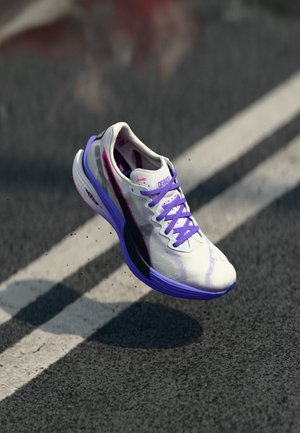 Zapatilla de running con parte superior en blanco y negro, acentos en púrpura y material texturizado. Presenta un diseño ligero y una suela prominente.