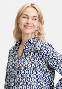 Hosszú ujjú blúz, amely kék, fehér és barna körmintával rendelkezik selymes anyagon, gallérral és gombos előlappal.