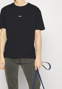 Person med svart T-shirt med "BOSS"-logotyp och grå byxor, som håller en blå läderrem eller hundkoppel i höger hand.