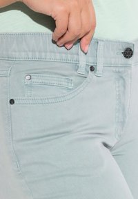 Jeans de mezclilla verde menta claro con una textura suave, que cuentan con un bolsillo frontal, costuras en contraste y un cierre de botón negro.
