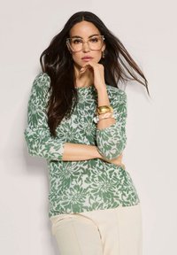 Frau mit langen, dunklen Haaren, die große Brille trägt, in einem grünen, floralen Pullover, beigen Hosen und goldenen sowie klaren Armreifen, posiert vor einer weißen Wand.