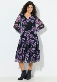 Frau mit lockigem Haar, die ein schwarzes Midikleid mit lila Blumenmuster und transparenten langen Ärmeln trägt, kombiniert mit schwarzen Stiefeletten, steht mit den Händen hinter dem Rücken.