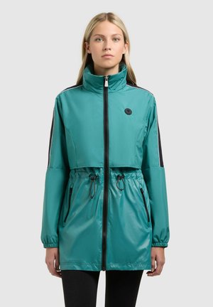 Młoda kobieta ubrana w turkusową kurtkę na zamek z wysokim kołnierzem, czarne paski na rękawach i ściągaczem w talii, stojąca na jednolitym tle.