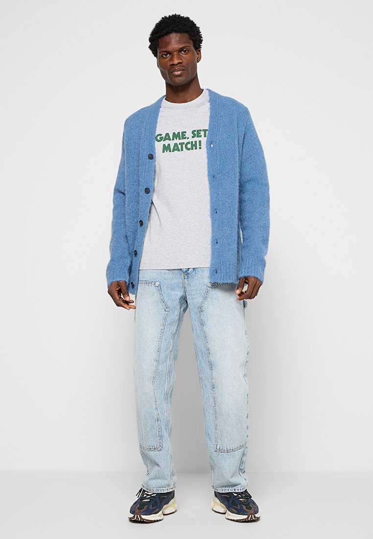 Blau gestrickte Strickjacke mit Knöpfen, graues T-Shirt mit grüner Schrift, hellblaue Jeans und gemusterte Sneaker.