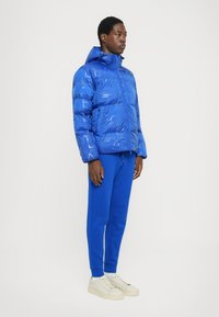 Chaqueta de plumas azul con un acabado texturizado y capucha, combinada con pantalones de chándal azules a juego y zapatillas de color claro.