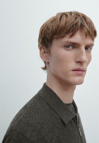 Maglione con zip grigio scuro e colletto, tessuto a trama, indossato sopra una maglia nera. Presenta un taglio di capelli corto e uno sfondo neutro.