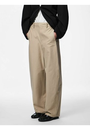 Persona con pantalones beige de pierna ancha, mocasines negros y un blazer negro con las manos en los bolsillos, de pie contra un fondo blanco.