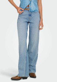 Femme portant un jean taille haute bleu clair à jambes larges, des bottes marron à bouts pointus, et un gilet en denim sans manches avec des boutons.