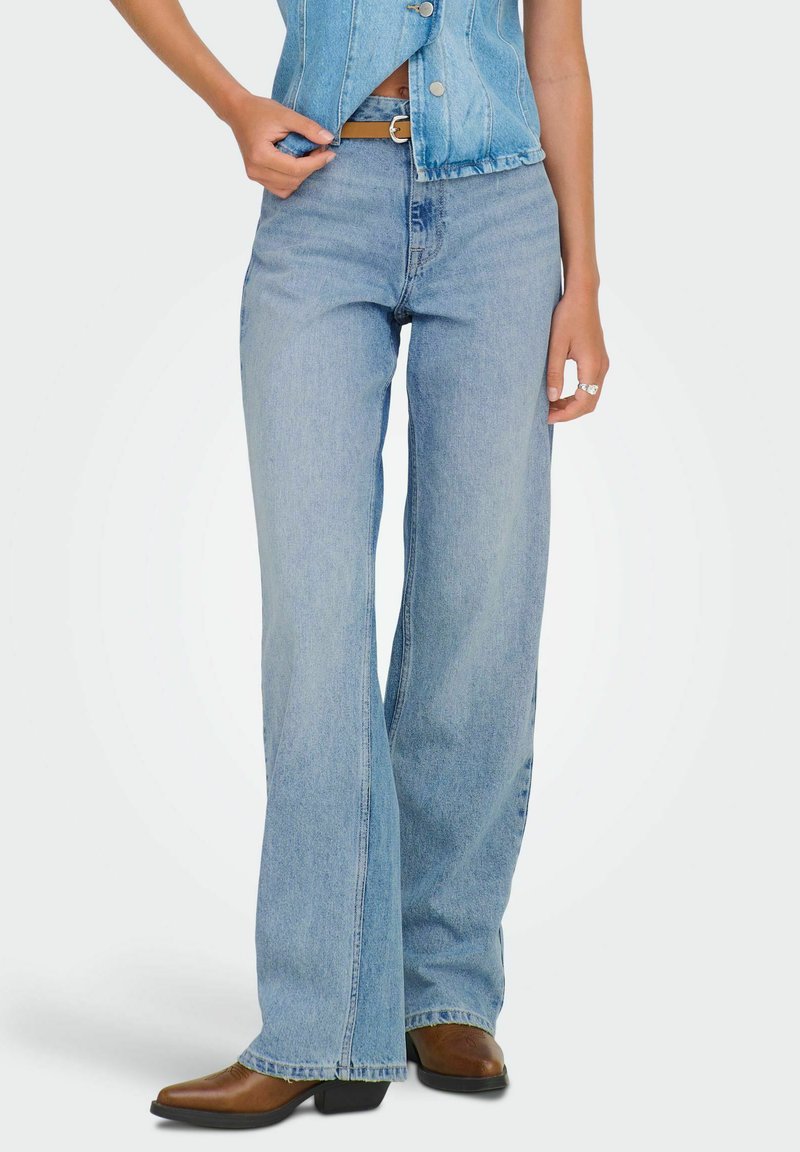 Femme portant un jean taille haute bleu clair à jambes larges, des bottes marron à bouts pointus, et un gilet en denim sans manches avec des boutons.