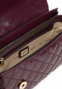 Sac à main en cuir matelassé violet avec intérieur beige, poche zippée et fermoir doré, marqué Guess Est. 1981 à l'intérieur.