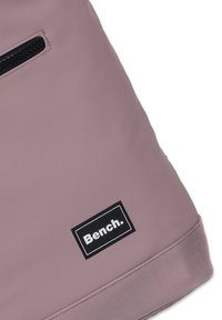 Sac rose clair avec une texture lisse, doté d'une poche zippée sur le côté et d'un logo rectangulaire noir "Bench." Design simple et moderne.