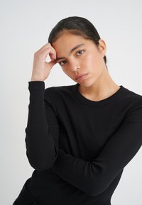 Svart ribbad långärmad topp med rund halsringning. Tyget verkar slätt med en figursydd design som visar upp texturerade detaljer på ärmarna.