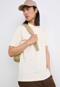 Polo shirt beige chiaro con maniche corte, dotato di colletto e tre bottoni, indossato con un cappellino beige e una tracolla beige dello zaino.