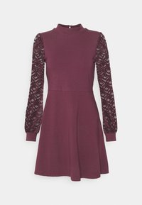 Lila klänning med hög hals, åtsittande överdel och utsvängd kjol. Långa ärmar har texturerad svart spets och rynkade detaljer vid axlarna.