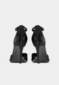 Ζευγάρι μαύρα πέδιλα με ψηλό τακούνι στιλέτο, λουριά στον αστράγαλο, φωτογραφημένα από πίσω σε λευκό φόντο.