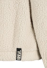 Texturiertes cremfarbenes Fleecegewebe mit einer weichen, flauschigen Oberfläche. Verfügt über ein schwarzes Etikett mit "SBWL" am Rand.