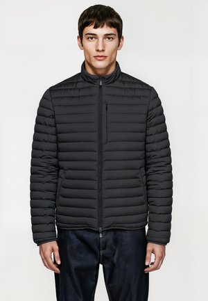 Zwarte pufferjack met een ritssluiting aan de voorkant, hoge kraag en horizontale stiksel. Bevat een klein ritszakje op de borst. Draagt over donkerblauwe jeans.