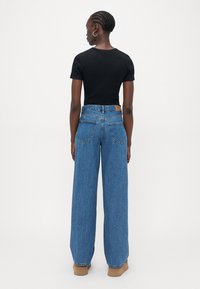 Bredde jeans i blå denim med høy midje, med to baklommer, et brunt lærmerke og en glatt, strukturert overflate. Svart tettsittende T-skjorte.