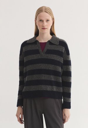 Femme aux longs cheveux blonds et raides, portant un pull à larges rayures gris foncé et noir et un pantalon gris foncé, debout devant un fond uni.