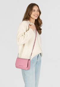 Bolso cruzado de cuero rosa con textura suave, herrajes plateados y una correa estampada, mostrado llevado sobre un suéter crema y unos jeans de mezclilla.