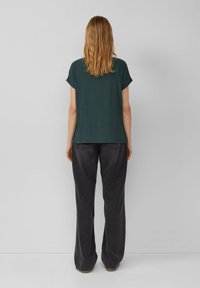 Donkergroen top met korte mouwen en een textuurpatroon, gecombineerd met hoge, wijde, donkergrijze broek, gezien vanuit de achterkant.