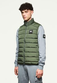 Oliivinvihreä täytetty liivi jossa on korkea kaulus, etuvetoketju ja musta logopatch. Käytetään vaaleanharmaan pitkähihaisen paidan päällä, jossa on elastiset hihansuut.