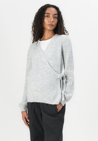 Lichtgrijze wikkeltrui van ribgebreid materiaal; heeft een striksluiting bij de taille en ontspannen mouwen. Gecombineerd met zwarte broek.